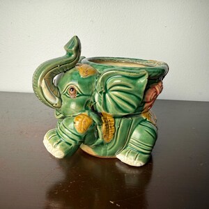 May include: A green ceramic elephant planter with a raised trunk and detailed features. The planter has a wide opening for plants and is decorated with yellow and pink accents. The elephant is sitting on its legs.