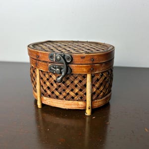 May include: A small, round, woven wicker box with a hinged lid and a metal latch. The box is brown and has a natural wood finish. It is sitting on a dark brown surface.