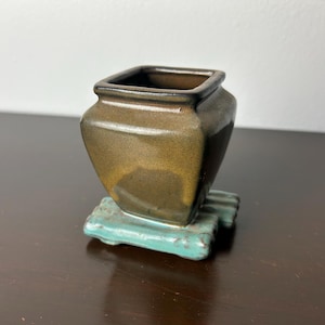 Puede incluir: Un pequeño macetero cuadrado de cerámica marrón con una base de color verde azulado. El macetero tiene una superficie ligeramente texturizada y un fondo redondeado.
