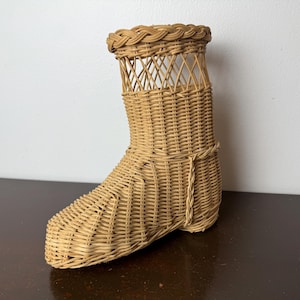 May include: A woven wicker boot-shaped basket in a natural tan color. The basket has a braided trim around the top and a decorative openwork design on the upper portion. The boot is displayed on a dark brown surface.