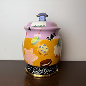 May include: A ceramic cookie jar with a lid, featuring a colorful design of cookies and other treats. The jar has a yellow and purple body with a black base and the word "Cookies" written on it.
