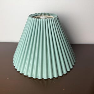 May include: A pleated lampshade in a soft, muted teal color. The shade has a classic, conical shape with vertical pleats and a scalloped edge. The lampshade is designed to diffuse light and add a decorative element to a lamp.