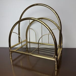 May include: A gold-colored metal magazine rack with an arched design. The rack has a rectangular base with vertical dividers and a series of curved bars forming the top. The metal has a shiny finish.