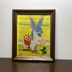 K&ouml;nnte beinhalten: Ein gerahmtes Filz-Applikationsbild mit einem Esel, der einen Korb mit Blumen tr&auml;gt. Der Esel ist hellgrau mit blauen Ohren und einer rosa Nase. Der Hintergrund ist gelb mit Streifen. Das Kunstwerk ist signiert mit "Diana 1976".