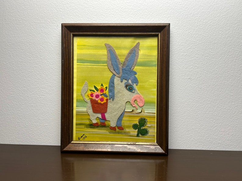 K&ouml;nnte beinhalten: Ein gerahmtes Filz-Applikationsbild mit einem Esel, der einen Korb mit Blumen tr&auml;gt. Der Esel ist hellgrau mit blauen Ohren und einer rosa Nase. Der Hintergrund ist gelb mit Streifen. Das Kunstwerk ist signiert mit "Diana 1976".