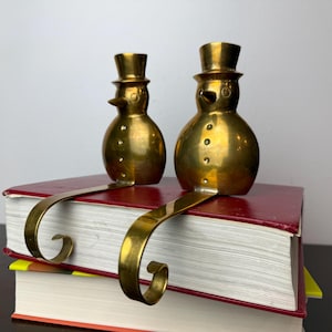 May include: Two brass snowman stocking holders with top hats and curved hooks. The snowmen have button details and are perched on a stack of books with red covers. The hooks are designed to hang stockings during the holiday season.