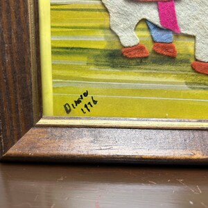 K&ouml;nnte beinhalten: Ein gerahmtes Kunstwerk mit gelbem und gr&uuml;nem Hintergrund. Das Kunstwerk ist aus Filz gefertigt und zeigt eine Szene mit zwei Figuren. Die Signatur "Dianne 1116" ist in der unteren rechten Ecke sichtbar.