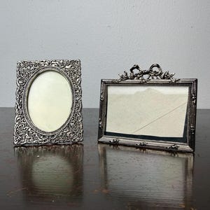 Könnte beinhalten: Zwei antike silberne Bilderrahmen. Einer ist rechteckig mit ovaler Öffnung und verschnörkelter Verzierung. Der andere ist rechteckig mit einer Schleife und floralem Design oben. Beide Rahmen haben einen hellfarbigen Innenraum.