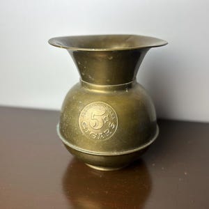 Puede incluir: Un jarrón de latón para cigarros de 5 centavos con una abertura ancha y acampanada. El jarrón es redondo y tiene un acabado liso y pulido.