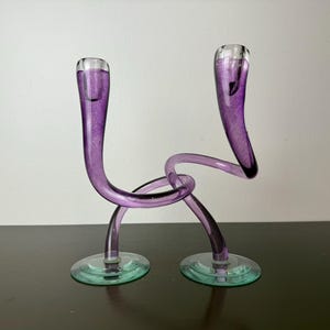May include: A pair of decorative glass candle holders. The holders are a vibrant purple color with clear tops and green bases. The stems are curved and intertwined, creating a unique and artistic design.