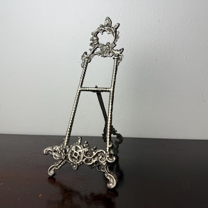 May include: A silver-toned metal display easel featuring an ornate, scrolled design at the top and base. The stand has two twisted vertical supports connected by a horizontal bar and a hinged rear leg for stability, creating a decorative frame.