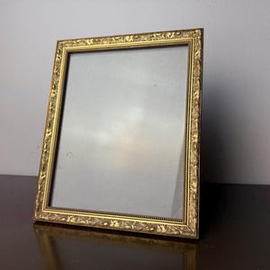 May include: A rectangular gold-colored picture frame with ornate floral detailing. The frame is angled slightly, showcasing its decorative border and the empty space within. The frame is resting on a dark brown surface.