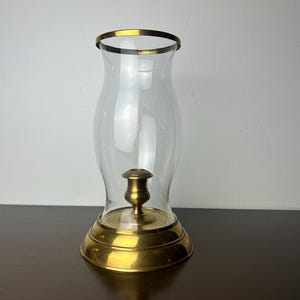 May include: A clear glass hurricane candle holder with a gold-colored rim and base. The candle holder has a brass-colored candle holder in the center. The candle holder is on a dark brown surface.