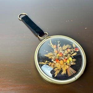 May include: A round, gold-framed picture with a black background and a dried flower arrangement inside. The flowers are a mix of yellow, orange, and red.