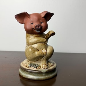Może przedstawiać: Ceramiczna figurka uśmiechniętej świni w pozycji siedzącej. Świnia ma brązową głowę i uszy oraz jasnozielone ciało. Figurka stoi na okrągłej podstawie z kwiatowym wzorem.