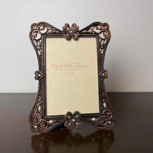 May include: A rectangular picture frame with a dark bronze finish, featuring ornate scrollwork, flower-shaped accents at the top and bottom, and embedded clear crystals and faux pearls. The insert reads Watch Hill Framers 4" x 6".
