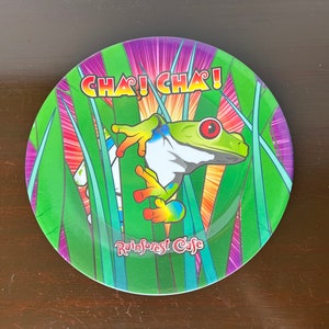 Può includere: Un piatto rotondo colorato con una rana verde su uno sfondo verde e viola. Il piatto ha le parole "Cha! Cha!" e "Rainforest Cafe" stampate su di esso.