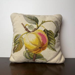 May include: Square decorative pillow with a needlepoint design of a yellow and pink peach with green leaves and a brown stem. The pillow has a cream-colored background and is placed on a dark brown surface.
