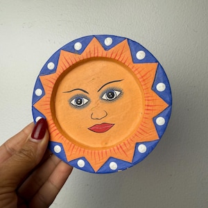 May include: A circular painted plate featuring a central orange sun face with detailed eyes, eyebrows, and red lips. The border is a blue ring with orange sun rays and white polka dots. The design is hand-painted with a matte finish on a flat surface.