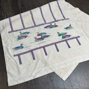 May include: White bath towel with a blue, red, and green embroidered design of ducks swimming on a pond. The towel has a striped border.