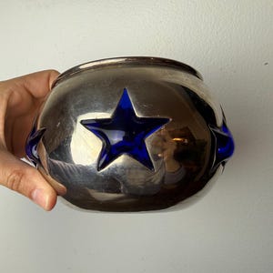 Pode incluir: Uma tigela redonda prateada com três recortes em forma de estrela azul. A tigela tem uma superfície refletora e uma borda lisa e arredondada. As estrelas azuis adicionam um toque de cor e contrastam com o acabamento metálico.