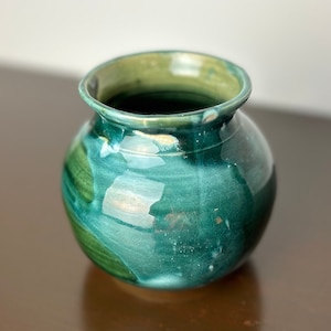 Könnte beinhalten: Eine kleine, runde Keramikvase mit einer grün-weißen Glasur. Die Vase hat eine breite Öffnung und einen abgerundeten Boden.