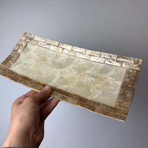 May include: Rectangular decorative tray with a mosaic border. The tray's centre has a pearlescent finish, and the border is made of small, rectangular tiles in shades of gold and brown. The tray is held against a plain, light grey background.
