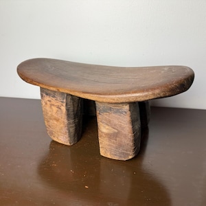Puede incluir: Un pequeño taburete de madera hecho a mano con un asiento curvo y tres patas resistentes. El taburete está hecho de madera de color marrón oscuro, que muestra la veta y la textura naturales. Es un mueble rústico.