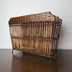 Puede incluir: Un revistero vintage hecho de bambú tejido y metal oscuro. El revistero tiene forma rectangular con listones verticales de bambú y una parte superior tejida. Las patas son delgadas y curvas. El revistero está diseñado para guardar revistas o periódicos.