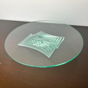 Puede incluir: Un juego de posavasos de vidrio transparente con forma curva, apilados uno encima del otro, sobre una bandeja redonda de vidrio transparente.