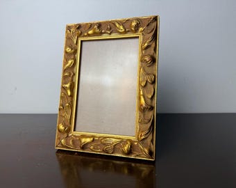 May include: A rectangular, ornate gold picture frame with a detailed floral and scroll design. The frame is angled, showcasing its intricate carvings and the empty space for a photograph or artwork. The frame is made of a gold-toned material.