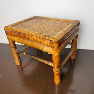 May include: A small, rectangular stool constructed from natural bamboo with a warm, mottled brown finish. The piece features a flat, slatted top surface supported by four sturdy bamboo legs connected by horizontal crossbars for structural stability.