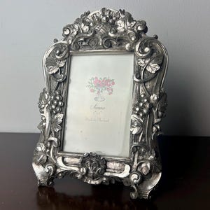 Puede incluir: Un marco de fotos plateado con un diseño ornamentado con vides y hojas. El marco tiene un paspartú blanco y una pequeña imagen de flores rosas en el interior. El marco dice "Sianna Made in Thailand".