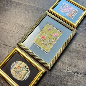 Könnte beinhalten: Drei goldgerahmte Bilder mit gestickten Blumendesigns und Vögeln. Die Bilder befinden sich auf einem Holzboden.