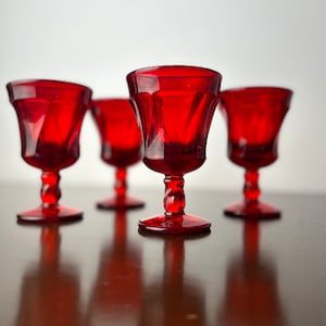 Puede incluir: Cuatro copas de vidrio rojo con un diseño acanalado se colocan sobre una superficie de madera. Las copas están dispuestas en fila, con las dos del medio ligeramente más juntas.