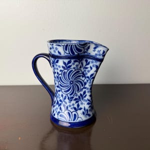 Puede incluir: Jarra de cerámica azul y blanca con un diseño floral. La jarra tiene un asa curva y un pico para verter. El patrón floral incluye grandes flores arremolinadas y pequeños detalles de hojas y flores.