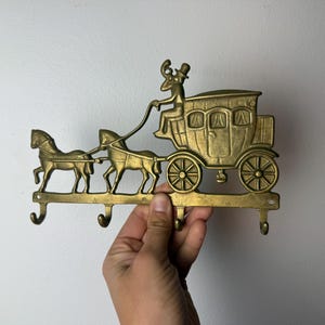 May include: Brass key holder featuring a horse-drawn carriage design. The carriage has detailed windows and wheels, with a driver figure. The key holder has four hooks for hanging keys. A decorative and functional item.