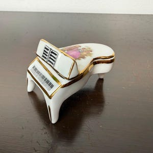 Op de afbeelding: Een wit porseleinen miniatuur vleugelpiano sieradendoosje met gouden rand. Het deksel is voorzien van een kleurrijke illustratie van twee figuren in een tuin. De piano heeft een zwart-wit toetsenbord en een bladmuziekstandaard.