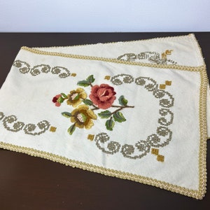 May include: A white embroidered table runner with a floral design featuring a pink rose and yellow flowers. The runner has a gold trim.
