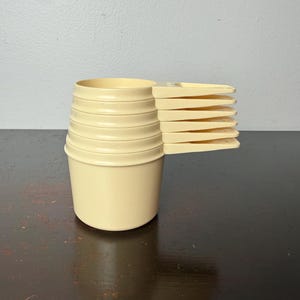 May include: A set of vintage cream-colored measuring cups and spoons. The cups are stacked, and the spoons are fanned out to the side. The set is made of a durable plastic material and is designed for kitchen use.