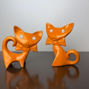 Puede incluir: Dos figuras de gato de cerámica naranja con ojos de pedrería y lazos. Un gato está de pie, el otro está sentado. Las figuras están sobre una superficie marrón oscuro.