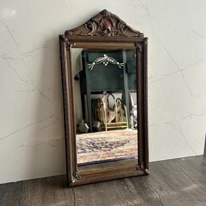 Puede incluir: Un espejo vintage con marco de madera tallado y un patrón floral grabado decorativo en el vidrio. El espejo tiene un acabado marrón oscuro y una forma rectangular.