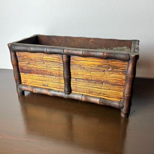 Puede incluir: Jardinera rectangular de bambú con marco y patas de color marrón oscuro. Los lados están hechos de tallos de bambú de color amarillo y marrón. La jardinera está diseñada para contener plantas y flores.