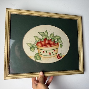 以下が含まれることがあります： イチゴの静物画が描かれた額入りのアートワーク。熟した赤いイチゴと緑の葉が詰まったバスケットが、濃い緑色の背景にクリーム色の楕円形に描かれています。フレームは金色です。