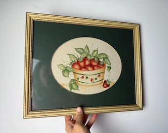 May include: Framed artwork featuring a still life of strawberries. The painting depicts a basket overflowing with ripe red strawberries and green leaves, set against a cream-colored oval on a dark green background. The frame is gold.