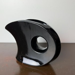 May include: A glossy black ceramic vase with a unique, abstract design. The vase features a large circular opening and curved, wing-like shapes. The smooth surface reflects light, highlighting its modern aesthetic. The vase is a decorative home accent.