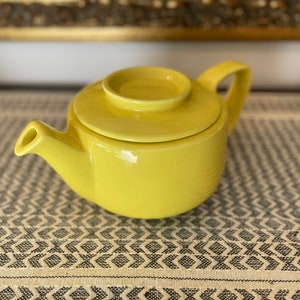 Op de afbeelding: Een felgele keramische theepot met een deksel. De theepot heeft een rond lichaam en een tuit aan de zijkant. De theepot staat op een gepatroneerd stofoppervlak.