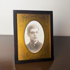 May include: An antique portrait in a rectangular black frame with a gold-colored oval interior. The portrait is a black and white image of a young man with glasses. The frame has a decorative floral design.