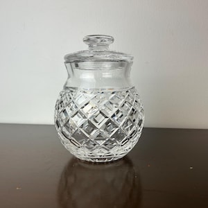 May include: A clear glass jar with a lid. The jar features a diamond-cut pattern on the lower half and a smooth upper half. The lid is also clear with a small knob. The jar is on a dark brown surface.
