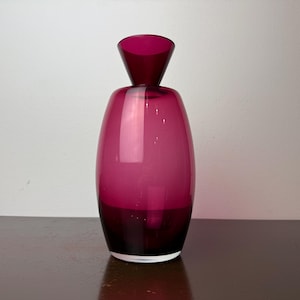 Könnte beinhalten: Eine hohe, elegante Vase aus magentafarbenem Glas. Die Vase hat einen abgerundeten Körper und einen ausgestellten, konischen Hals. Das Glas ist durchscheinend. Die Vase steht auf einer dunklen Oberfläche vor einer weißen Wand. Höhe ca. 25 cm.